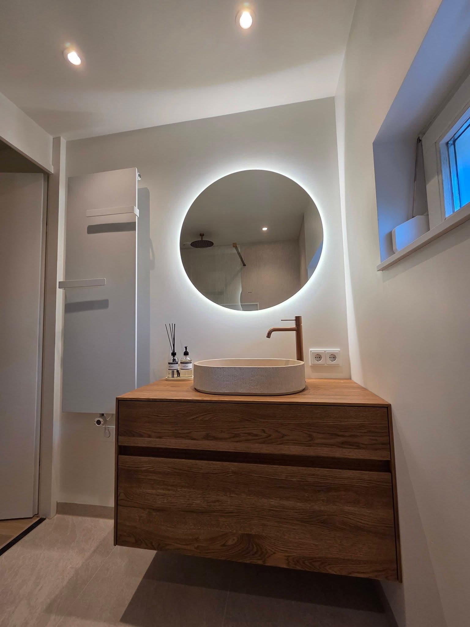 Badkamer met ronde verlichte spiegel en notenhouten wastafelmeubel